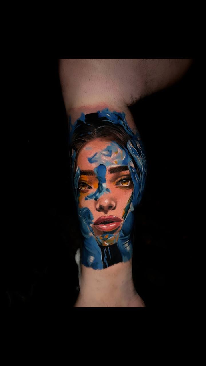 Abstraktes Tattoo eines Gesichts mit blauen Elementen; ausdrucksstarke Augen, realistisch gestaltet.