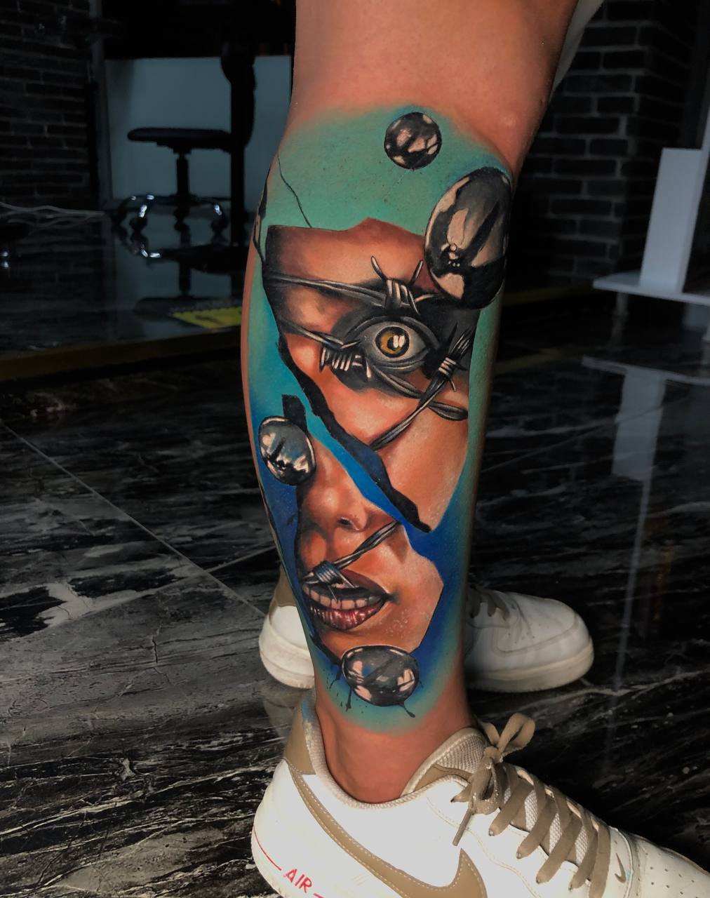 Realistisches Bein-Tattoo mit fragmentiertem Gesicht, blauem Hintergrund und metallischen Akzenten.