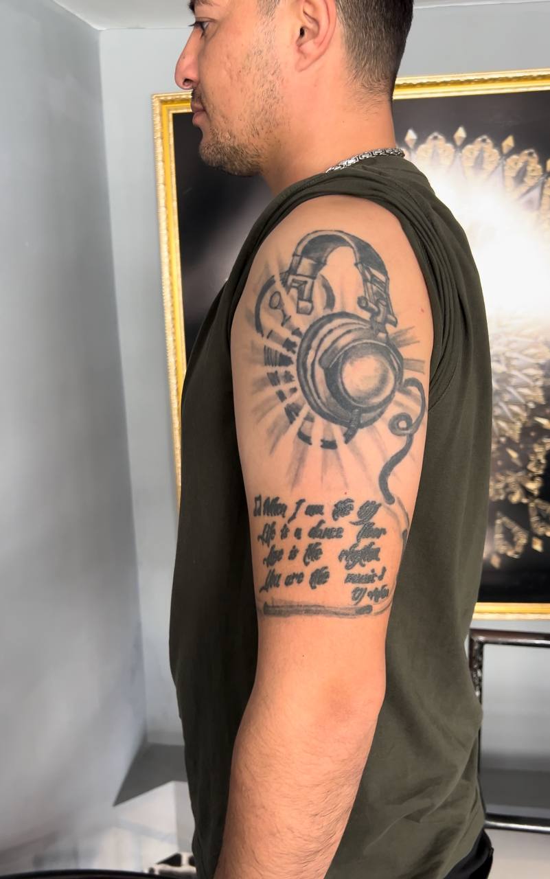 Mann in grünem Shirt zeigt Tattoo mit Kopfhörern und Zitat auf Oberarm, lächelt leicht.