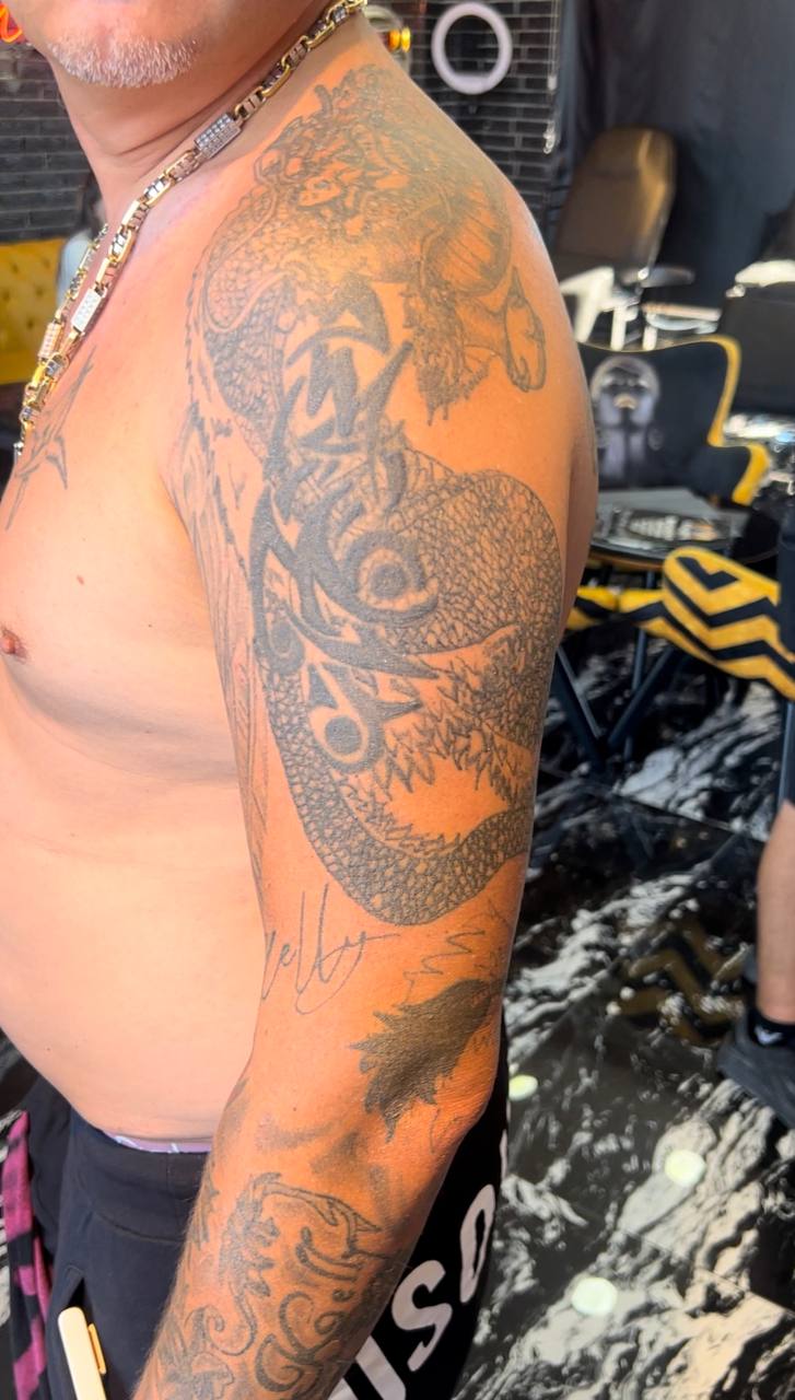 Mann mit Drachen-Tätowierung auf dem Arm, steht ohne Hemd. Hintergrund: Tätowierstube.