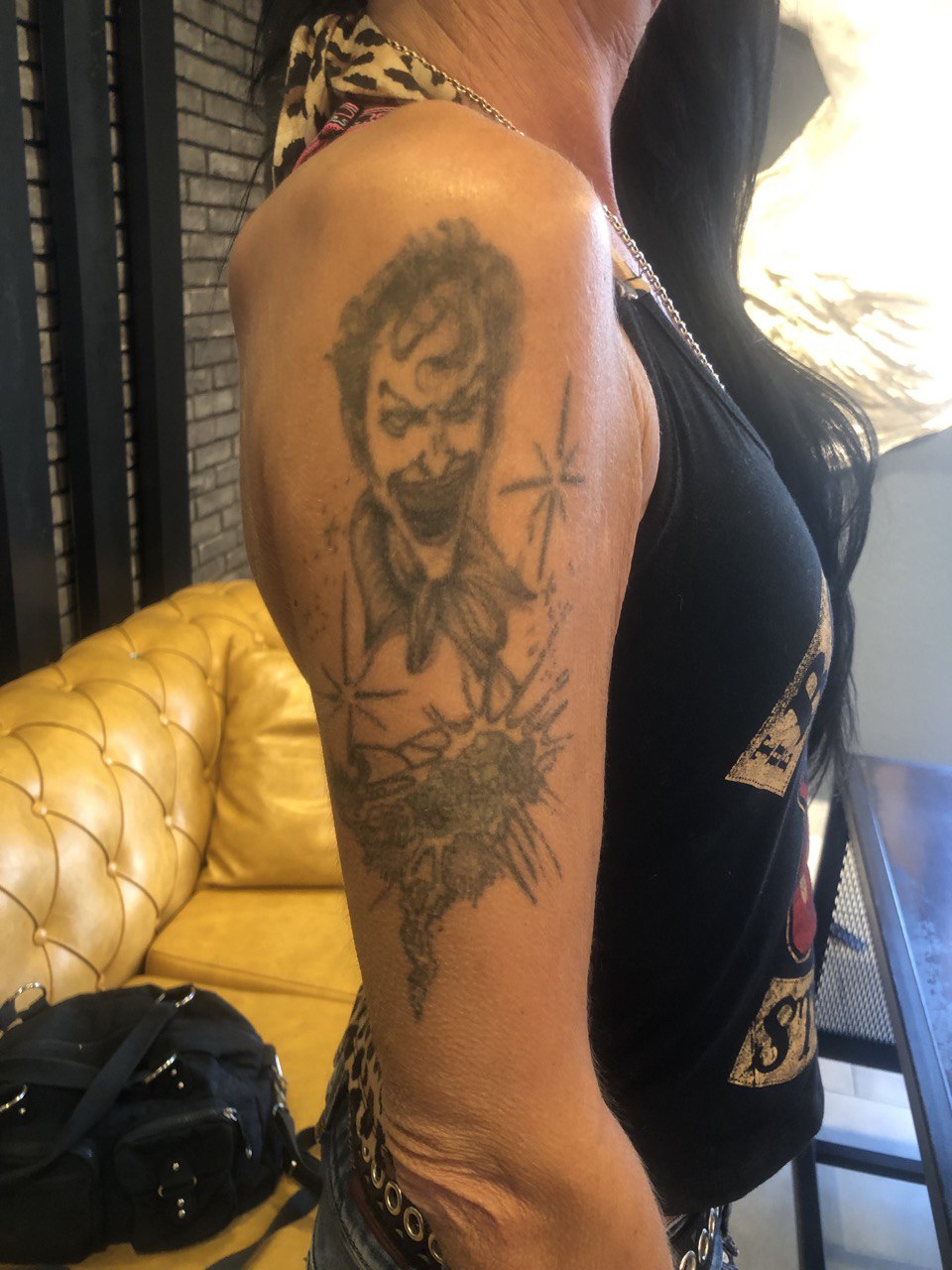 Frau zeigt Joker-Tattoo auf Oberarm, Lederjacke und Kette, gelbes Sofa im Hintergrund.
