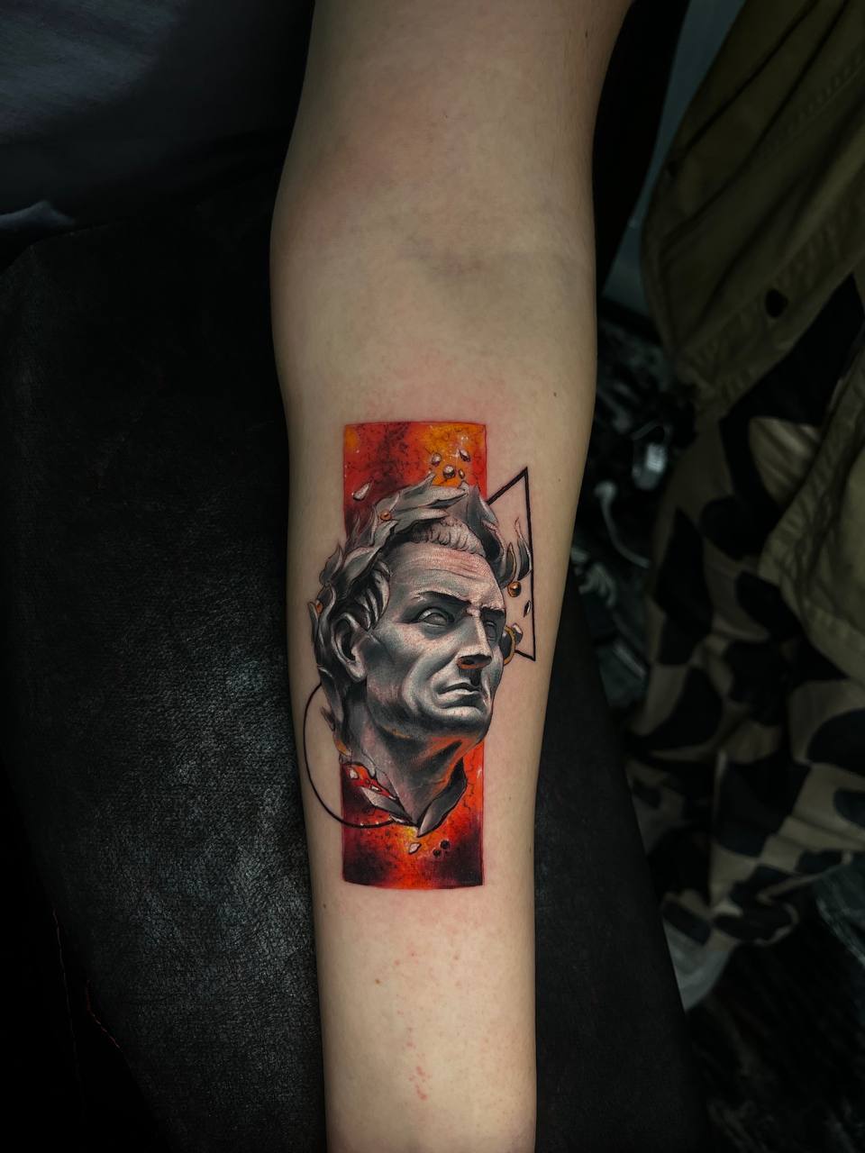 Tätowierung eines antiken Gesichts auf einem Arm, farbig, mit geometrischen Elementen.