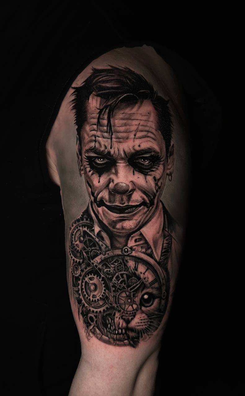 Tattoo eines grinsenden Mannes mit dunklem Haar und Uhrwerk-Elementen auf einem muskulösen Arm.