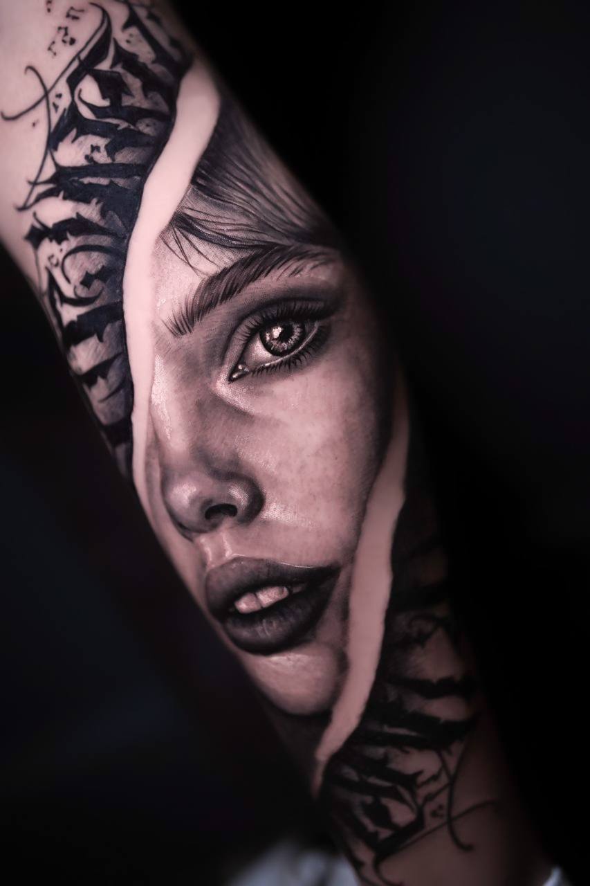Tattoo eines Gesichts mit intensiven Augen auf arm, dunkle Farbtöne, dynamische Linien.