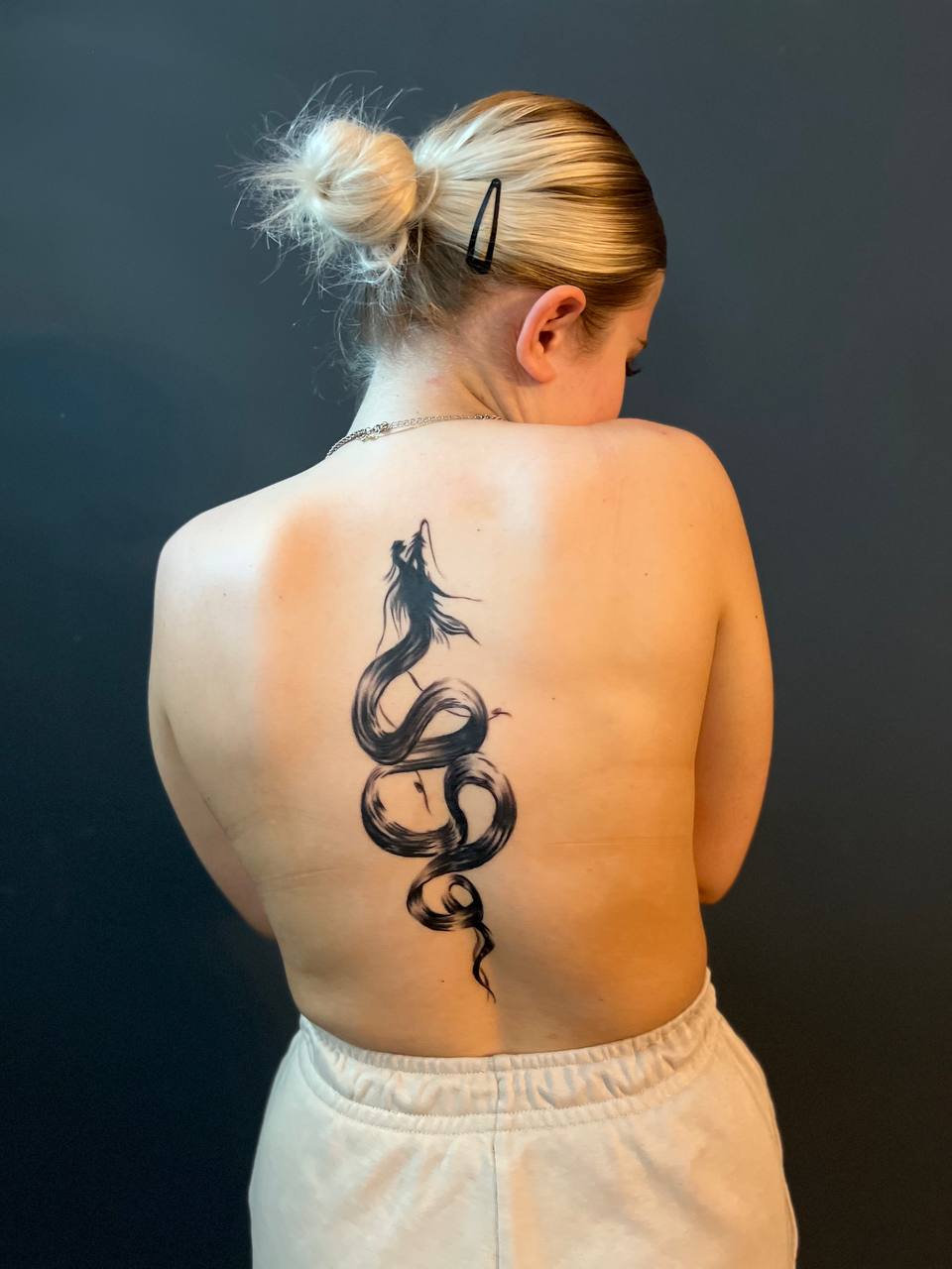Junge Frau mit Drachentattoo auf dem Rücken, auf dunkelgrauem Hintergrund, blonde Haare hochgesteckt.