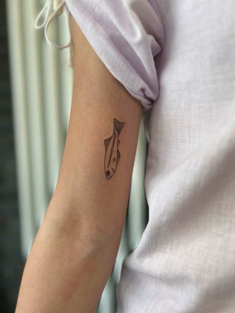 Fisch-Tattoo auf einem Arm in lila Shirt, neutraler Ausdruck, helle Haut.