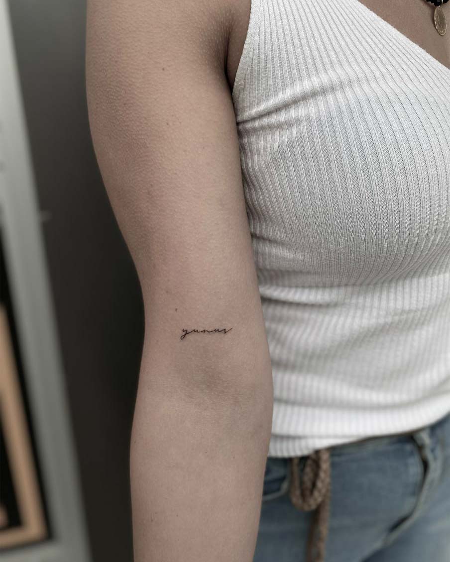 Person in weißem Top, zeigt kleinen Script-Tattoo auf dem Oberarm; Jeans tragen.