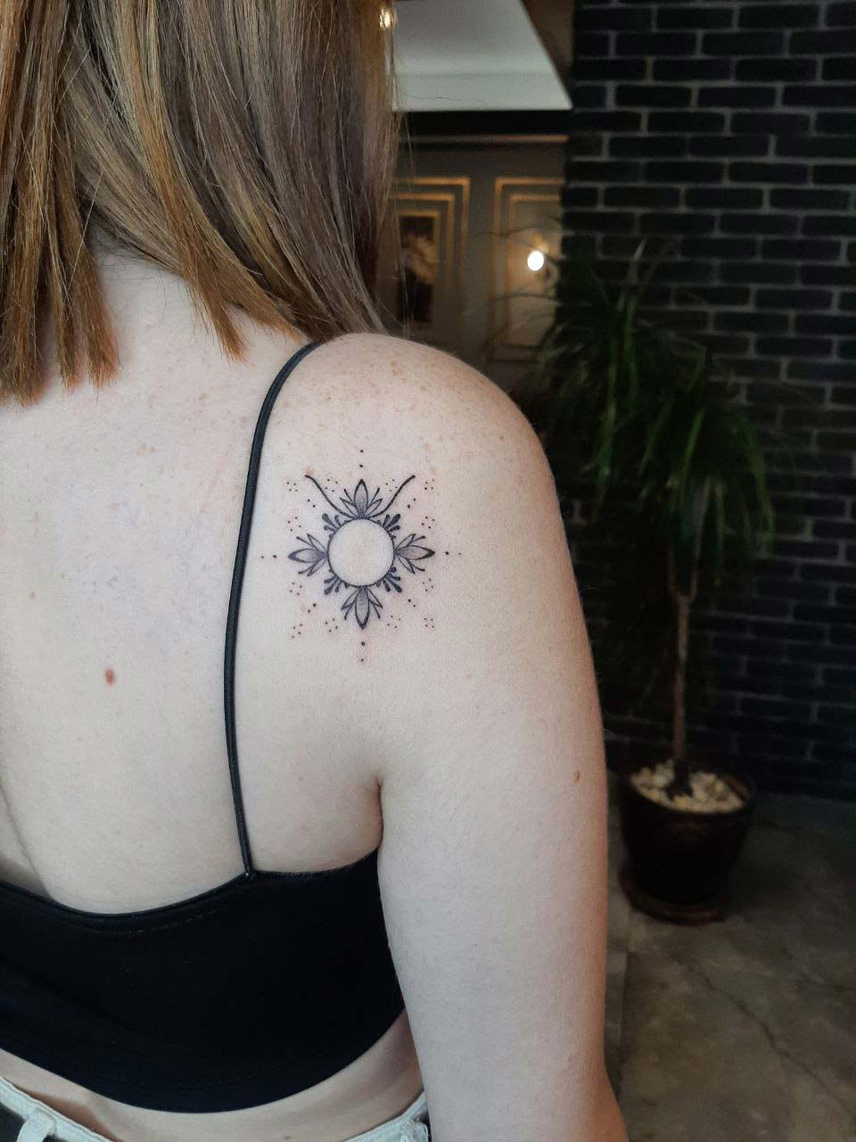 Frau mit Sonnen-Tattoo auf der Schulter, trägt schwarzes Top, braune Haare, entspannte Haltung.